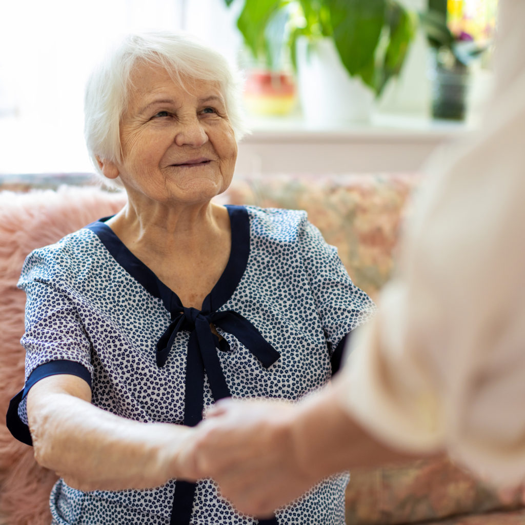 Female home carer supporting old woman to stand up from the sofa at care home